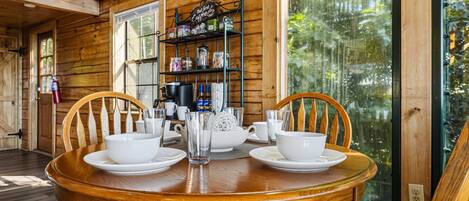 A wooden dining table with white plates and glasses, surrounded by chairs, in a cabin with a large window and shelves with kitchen items.