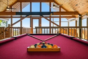 Ein Billardtisch mit Kugeln und Holzrahmen, platziert in einem Raum mit Holzbalken und großen Fenstern.