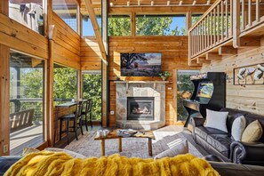 A cozy cabin interior with a stone fireplace, wooden walls, and a balcony with outdoor seating. - Mountain Top Cabin With Views Hot Tub and Arcade (Sevierville)