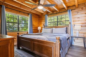 Ein Schlafzimmer einer Holzhütte mit einem Bett, einem Deckenventilator und großen Fenstern.