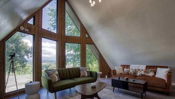 A spacious loft with large windows, a green sofa, a brown sofa, and a coffee table. - 4BR 3BA Cabin w Games Hot Tub Near Smoky Mtns (Kodak)