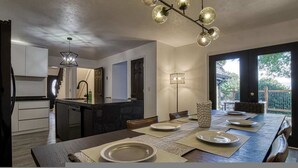 A modern kitchen with a central island, dining area with a long table set for four, and a view of the outdoors through a sliding door. - 4BR 3BA Cabin w Games Hot Tub Near Smoky Mtns (Kodak)