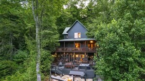 Eine zweigeschossige Holzhütte mit Terrasse und Feuerstelle, umgeben von dichtem Wald.