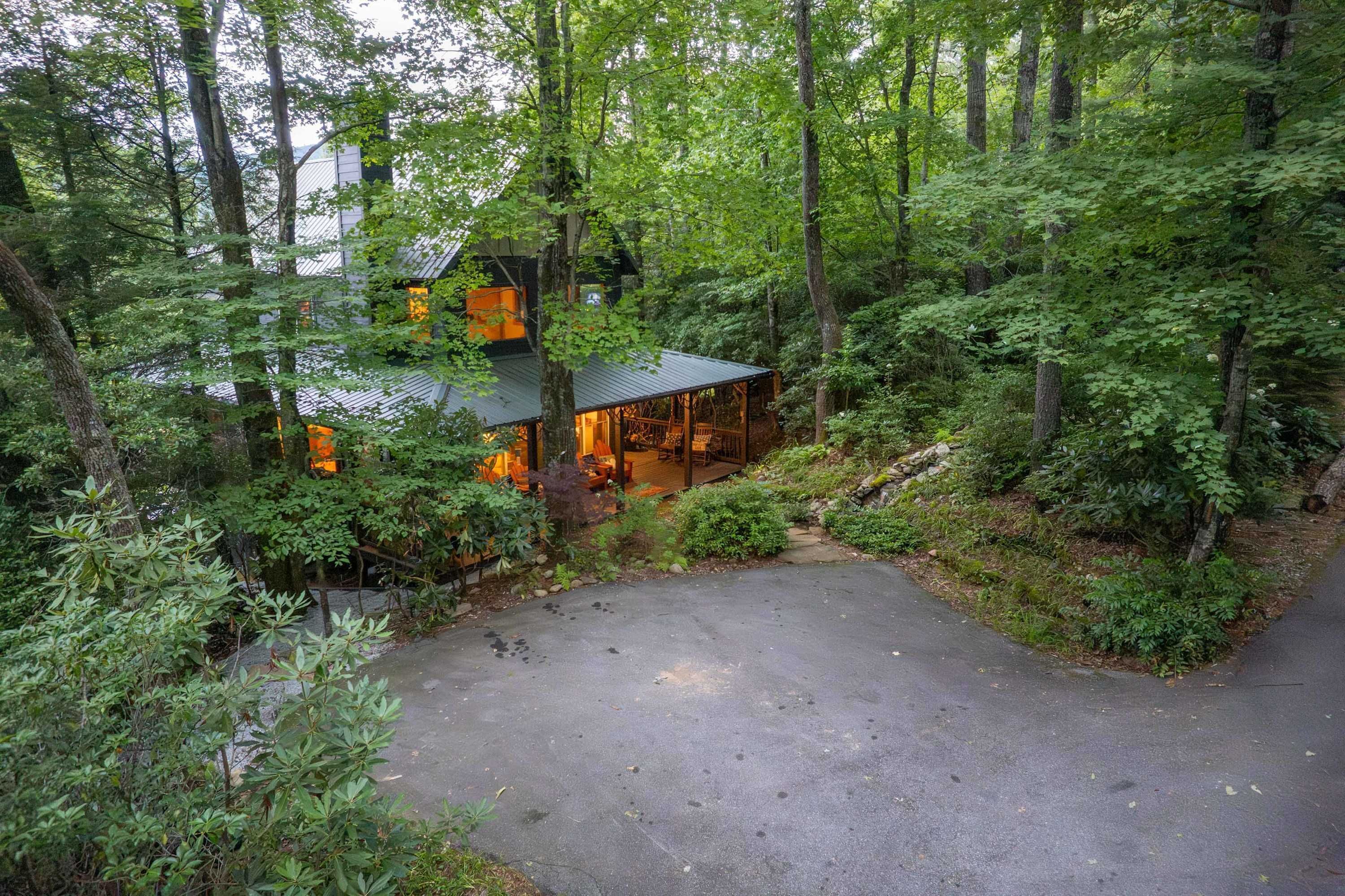 A secluded cabin surrounded by dense forest, with a paved driveway leading to a wooden deck.
