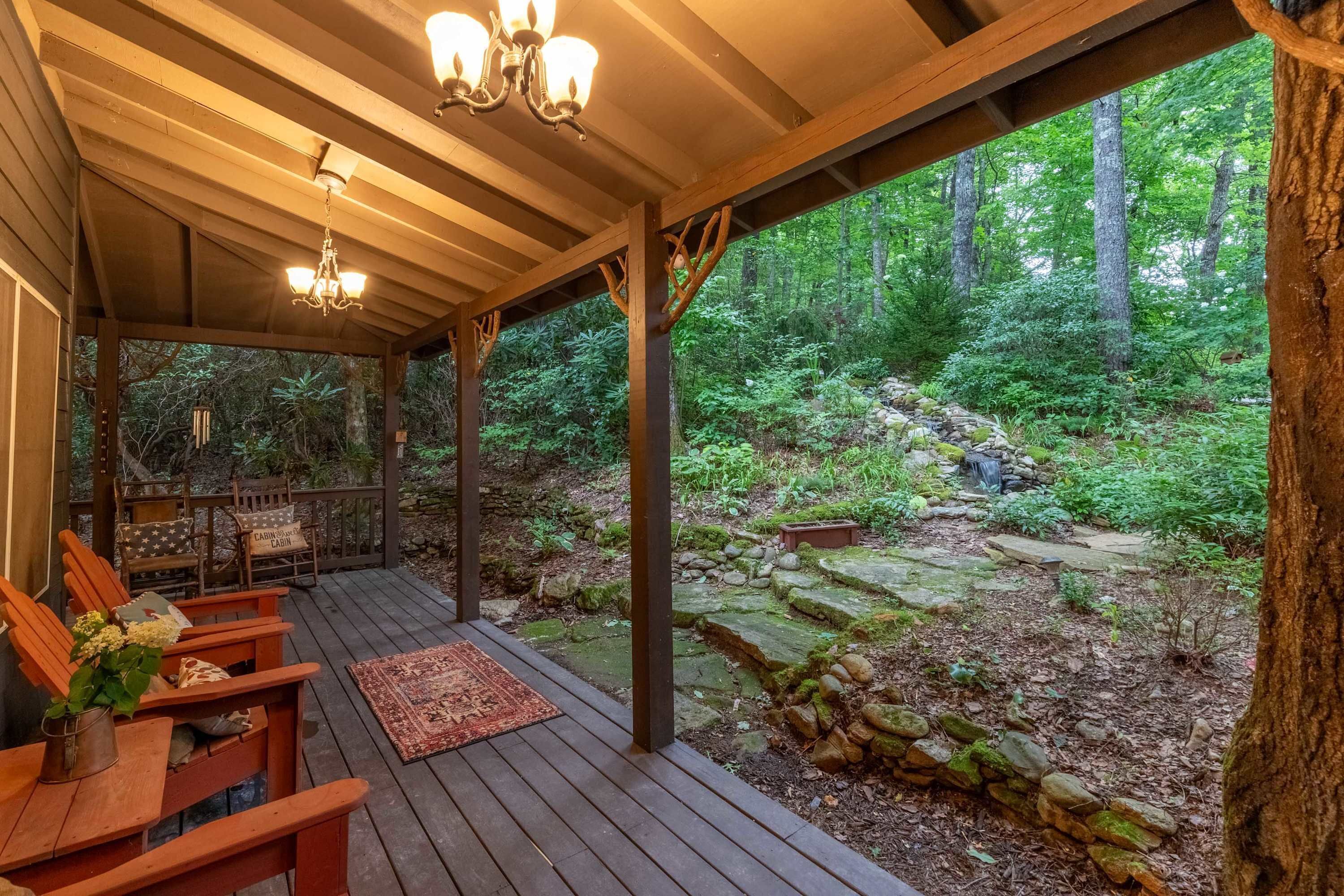 A wooden deck with benches and a rug, overlooking a forested area with a stone wall and a small waterfall.