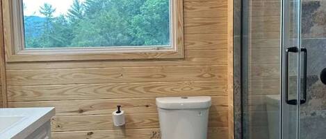 A bathroom with a wooden ceiling, a toilet, a glass shower enclosure, and a window with a view of trees.