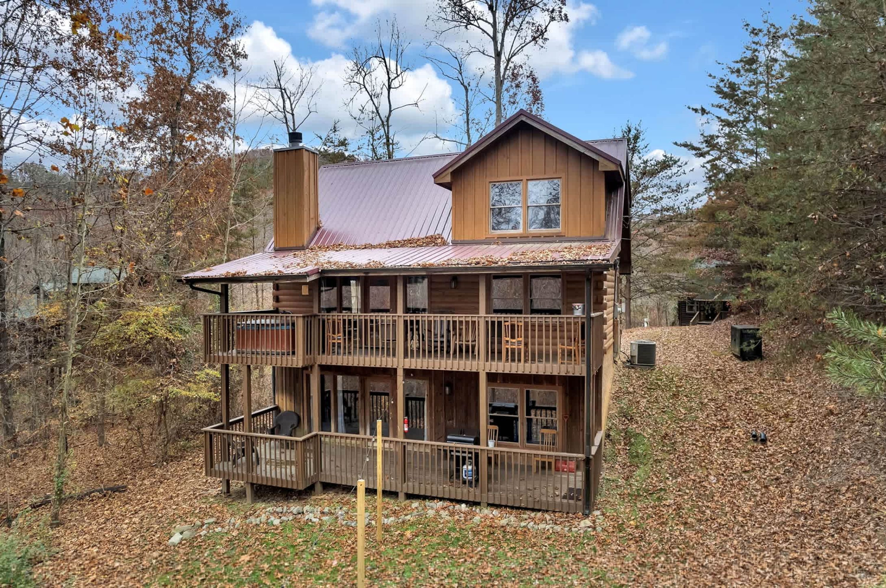 A two-story log cabin with a balcony, surrounded by trees and a metal roof.