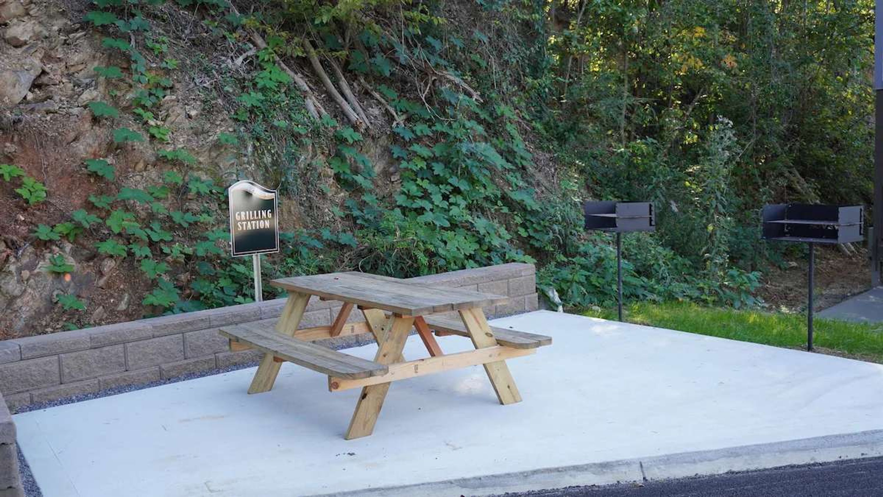 Une table de pique-nique avec une pancarte indiquant « Espace barbecue », devant un mur de pierres recouvert de verdure.