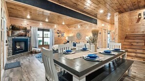 A cozy cabin interior with a stone fireplace, wooden walls, and a dining area with white chairs and blue plates. - Modern 5 Bedroom Cabin Close-dollywood Game Room (Pigeon Forge)