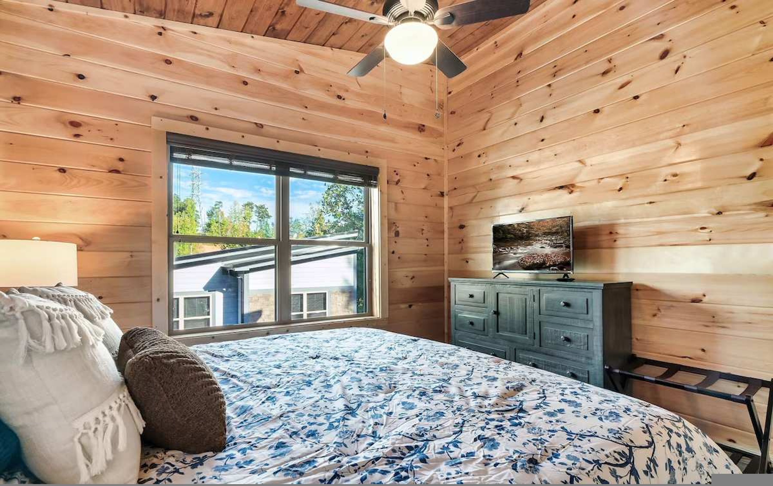 Une chambre avec une grande fenêtre, un lit recouvert d’une couvre-lit à motifs floraux, une commode et un ventilateur de plafond.