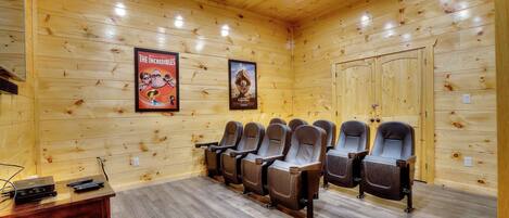A wooden-paneled room with a row of reclining chairs, a small table with electronic devices, and two posters on the wall.
