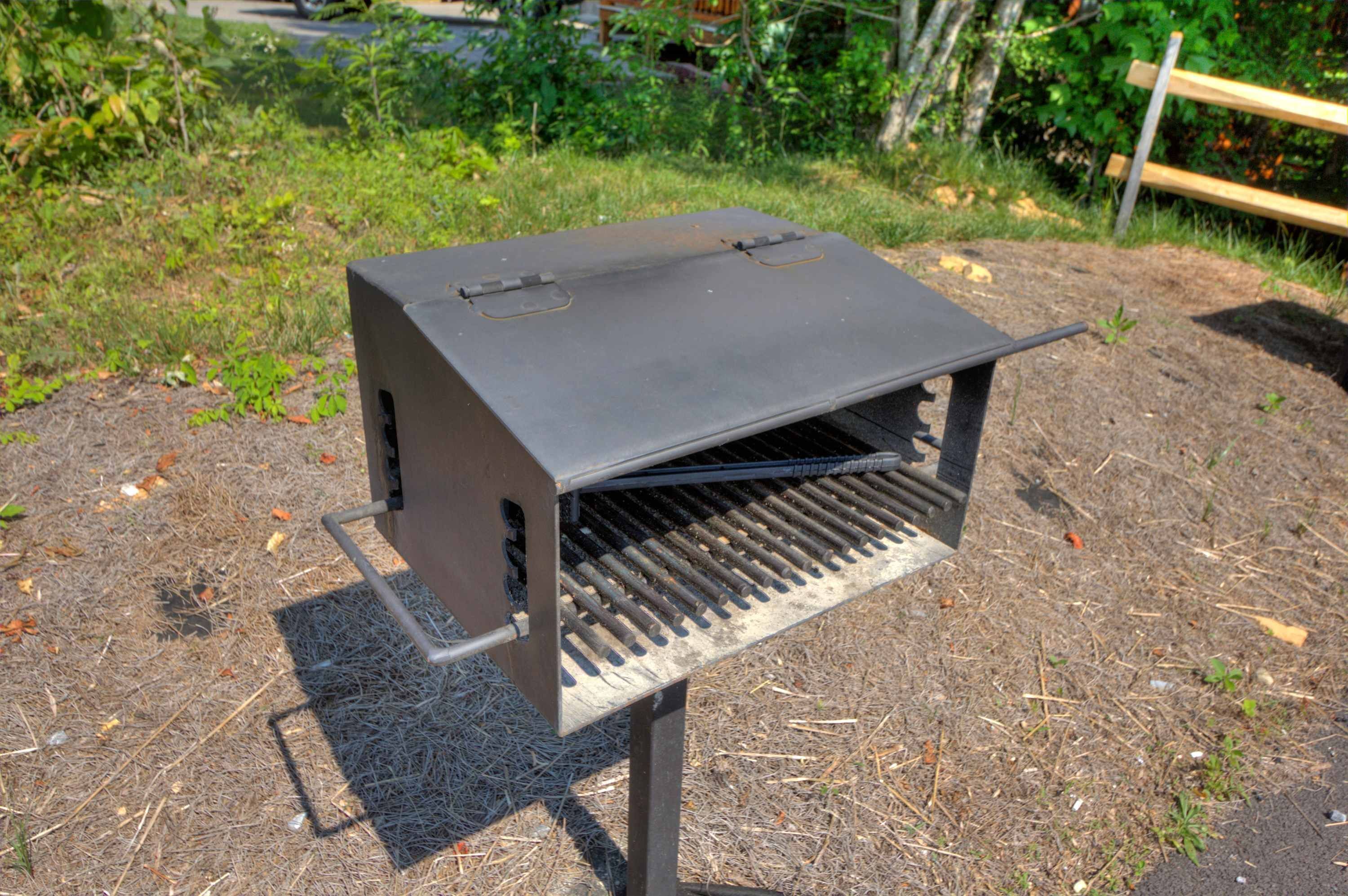 En grill med fladt grillområde og ventilationsgitter på siderne, placeret på en græsplæne med en træbænk i baggrunden.