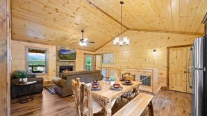 A wooden cabin interior with a dining table set for four, a sofa, a fireplace, and a kitchen area. - Stunning 4 King Suites Hot Tub Views Gamerm (Gatlinburg)