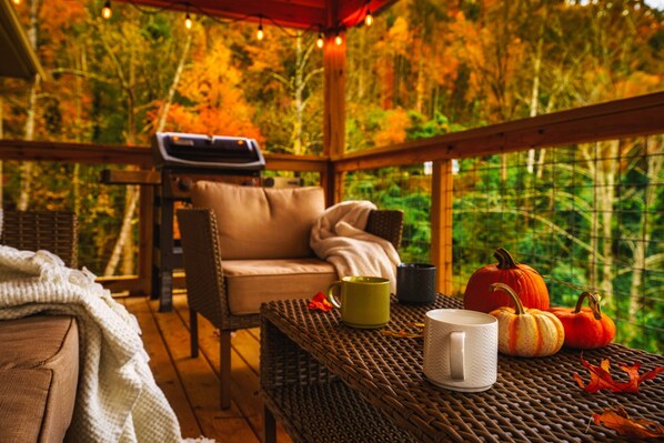 A cozy outdoor seating area with wicker furniture, a table with mugs and pumpkins, and a view of autumn foliage through a wooden railing. - Stunning 4 King Suites Hot Tub Views Gamerm (Gatlinburg)