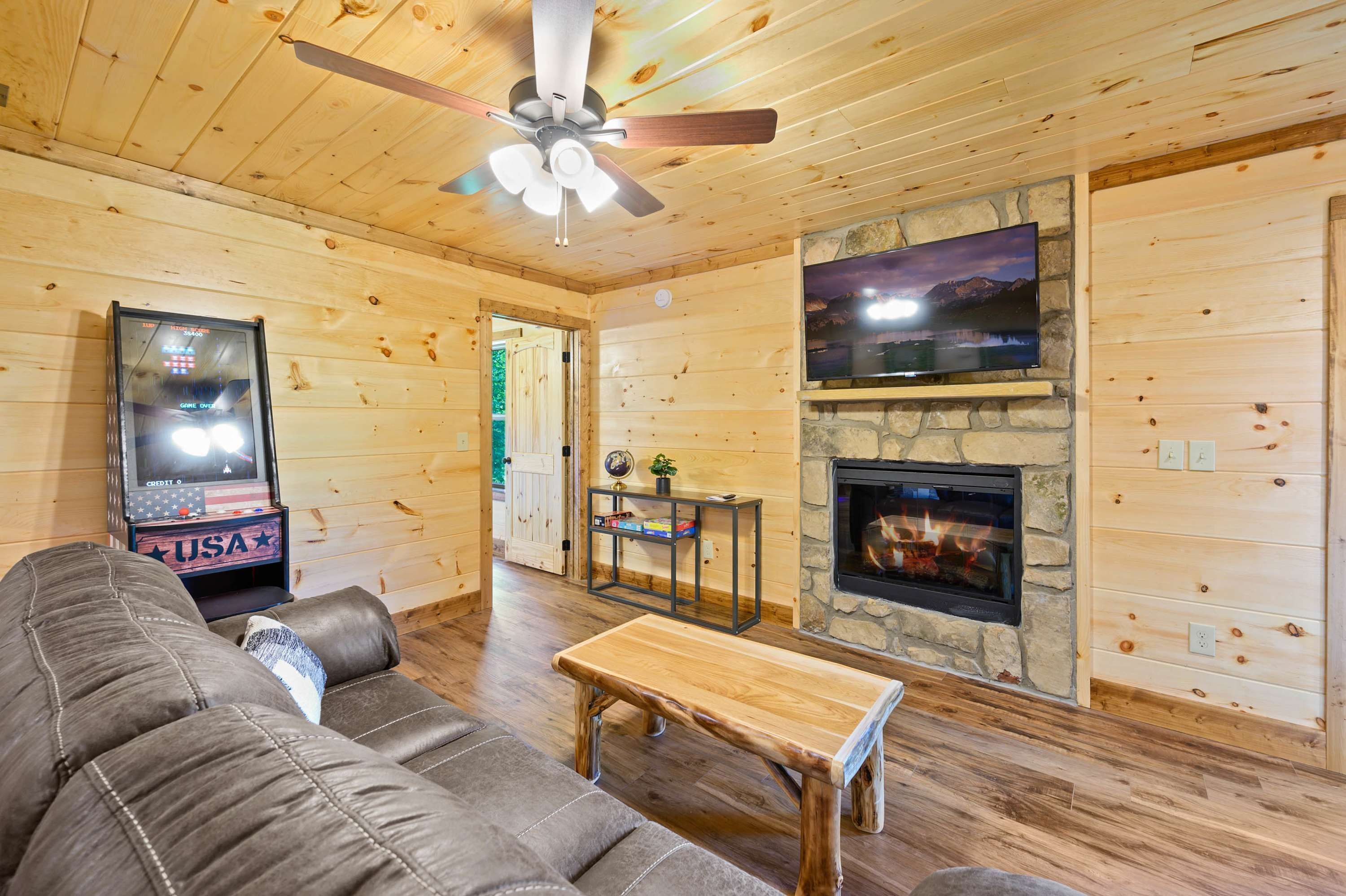 Une pièce confortable, aménagée dans le style d’un chalet, avec un plafond en bois, un ventilateur de plafond, une cheminée en pierre, un téléviseur fixé au mur en pierre, un canapé et une petite table en bois.