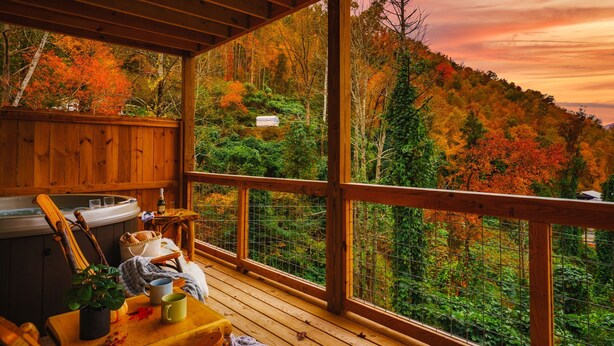 A wooden deck with a hot tub, a table with mugs, and a view of a forested hillside during autumn. - Stunning 4 King Suites Hot Tub Views Gamerm (Gatlinburg)