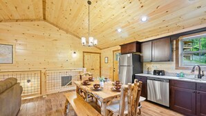 A wooden cabin interior with a dining table set for four, a kitchen with stainless steel appliances, and a window overlooking greenery. - Stunning 4 King Suites Hot Tub Views Gamerm (Gatlinburg)