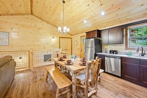 A wooden cabin interior with a dining table set for four, a kitchen with stainless steel appliances, and a window overlooking greenery. - Stunning 4 King Suites Hot Tub Views Gamerm (Gatlinburg)