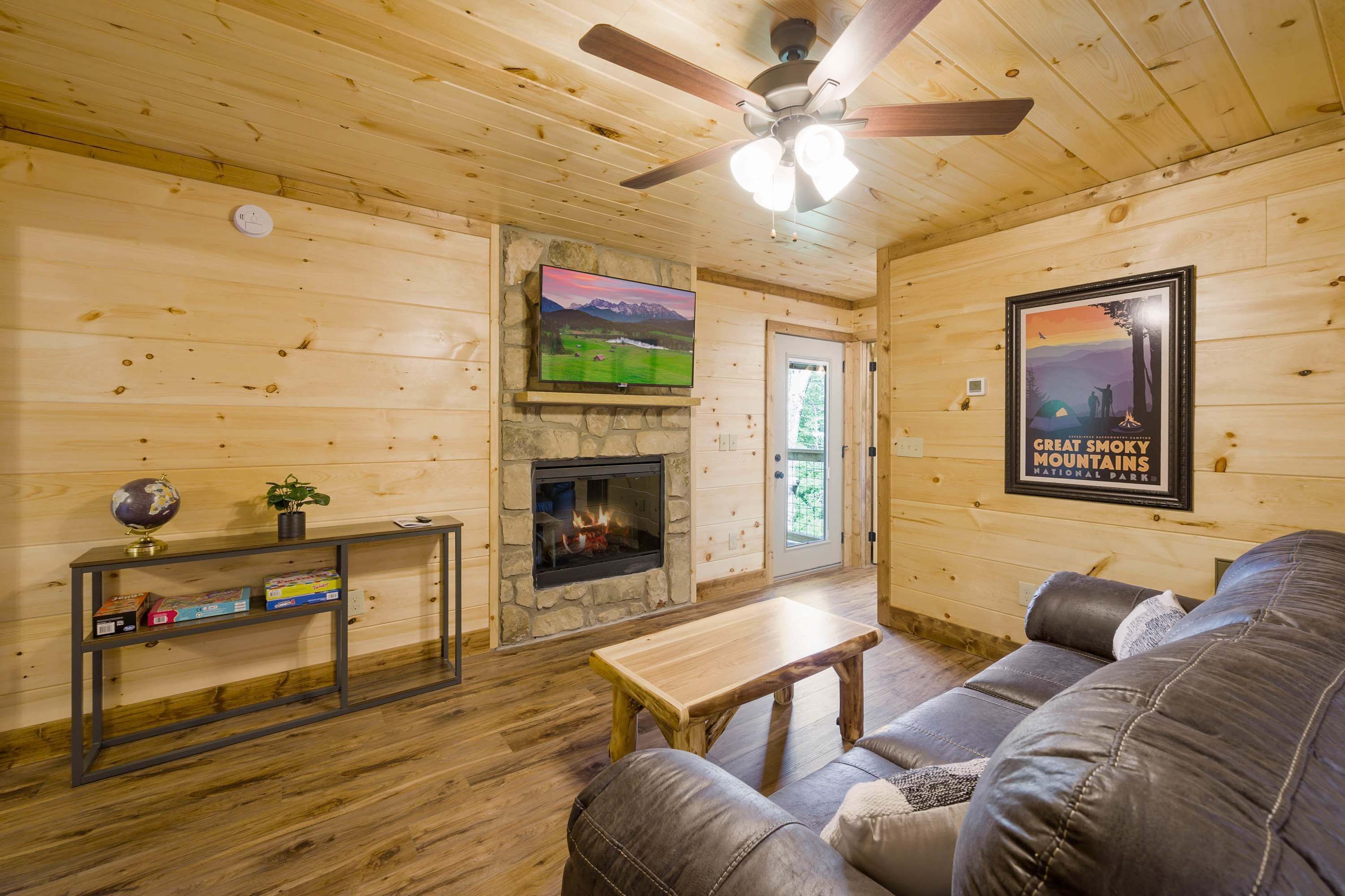 A cozy cabin interior with a stone fireplace, wooden walls, a leather couch, and a television mounted on the wall.