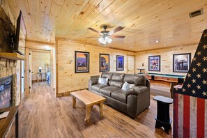 A wooden-floored room with a leather sofa, a wooden coffee table, a ceiling fan, and framed posters on the walls. - Stunning 4 King Suites Hot Tub Views Gamerm (Gatlinburg)