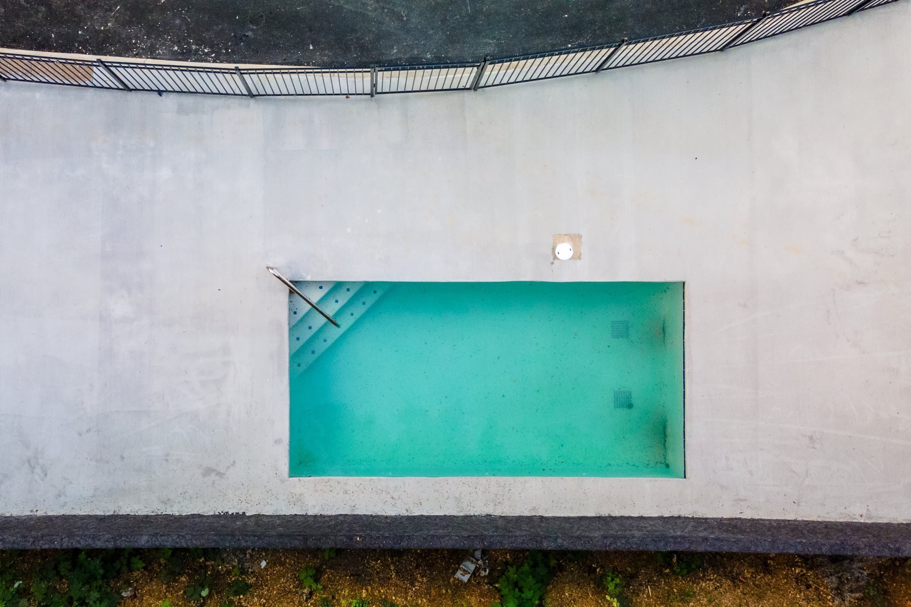 Piscina retangular com escada de metal e uma pequena janela, rodeada por um muro de concreto.