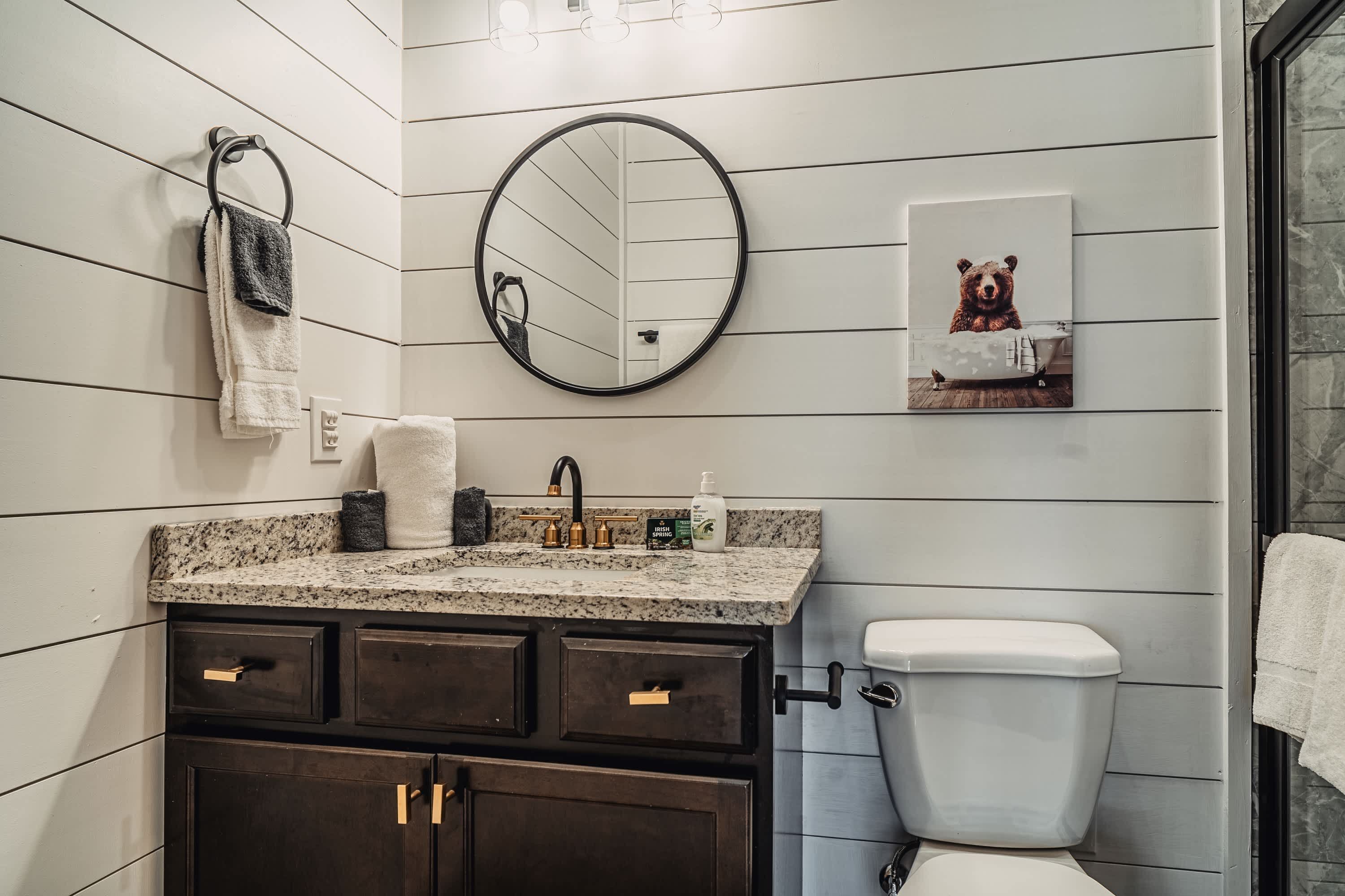 A bathroom with a granite countertop, dark wooden cabinets, a white toilet, a round mirror, and a bear-themed artwork on the wall.