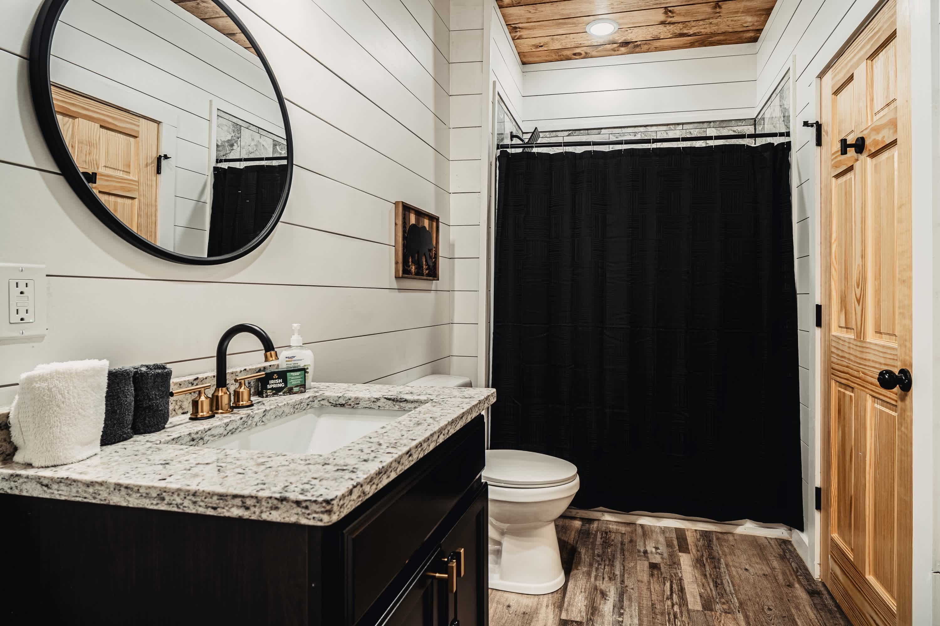 A bathroom with a white vanity, black shower curtain, and wooden flooring.