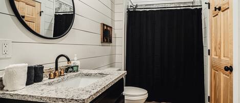 A bathroom with a white vanity, black shower curtain, and wooden flooring.