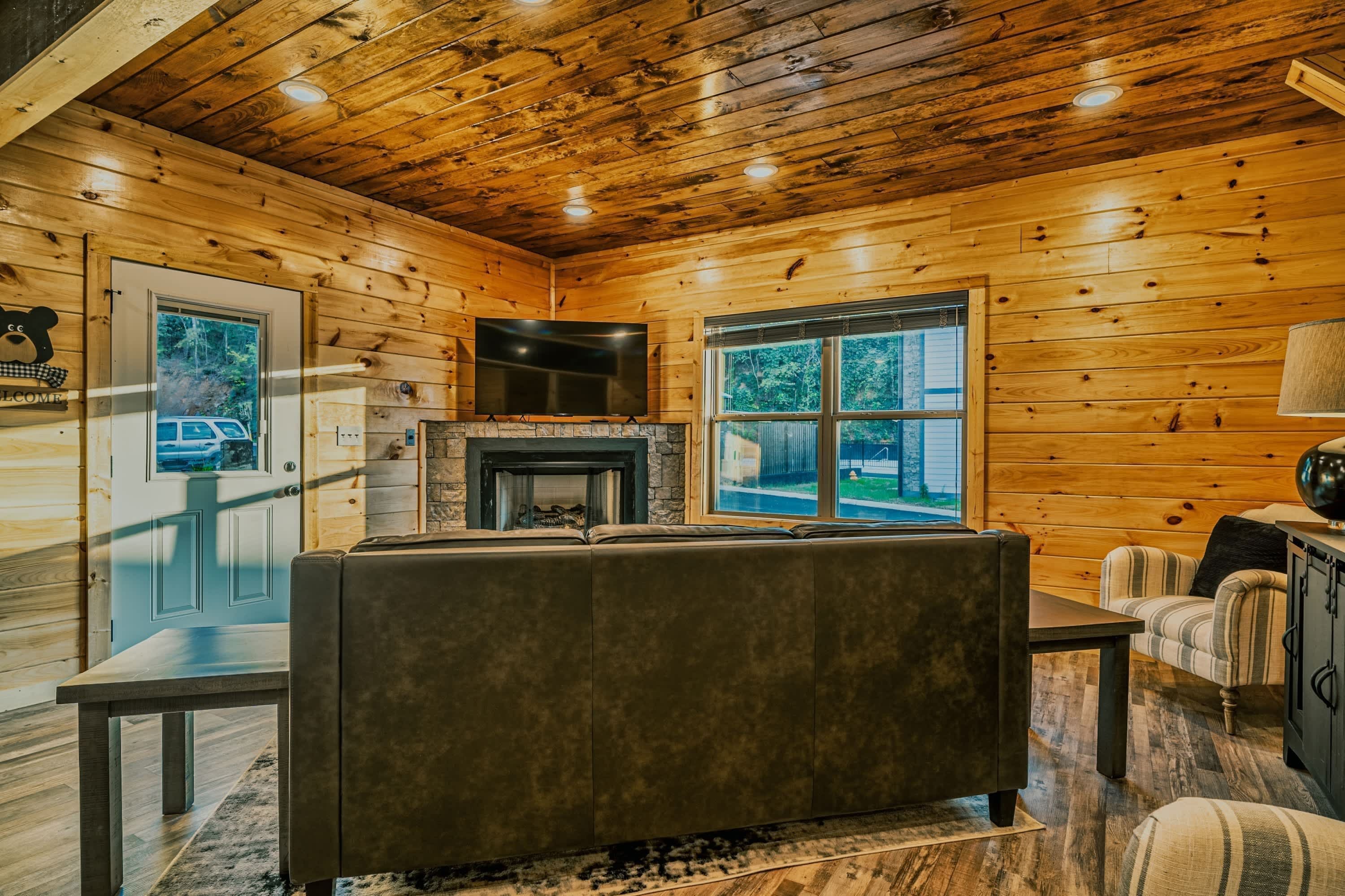 A log cabin interior with a stone fireplace, a sofa, a television mounted on the wall, and a window with a view of the outdoors.