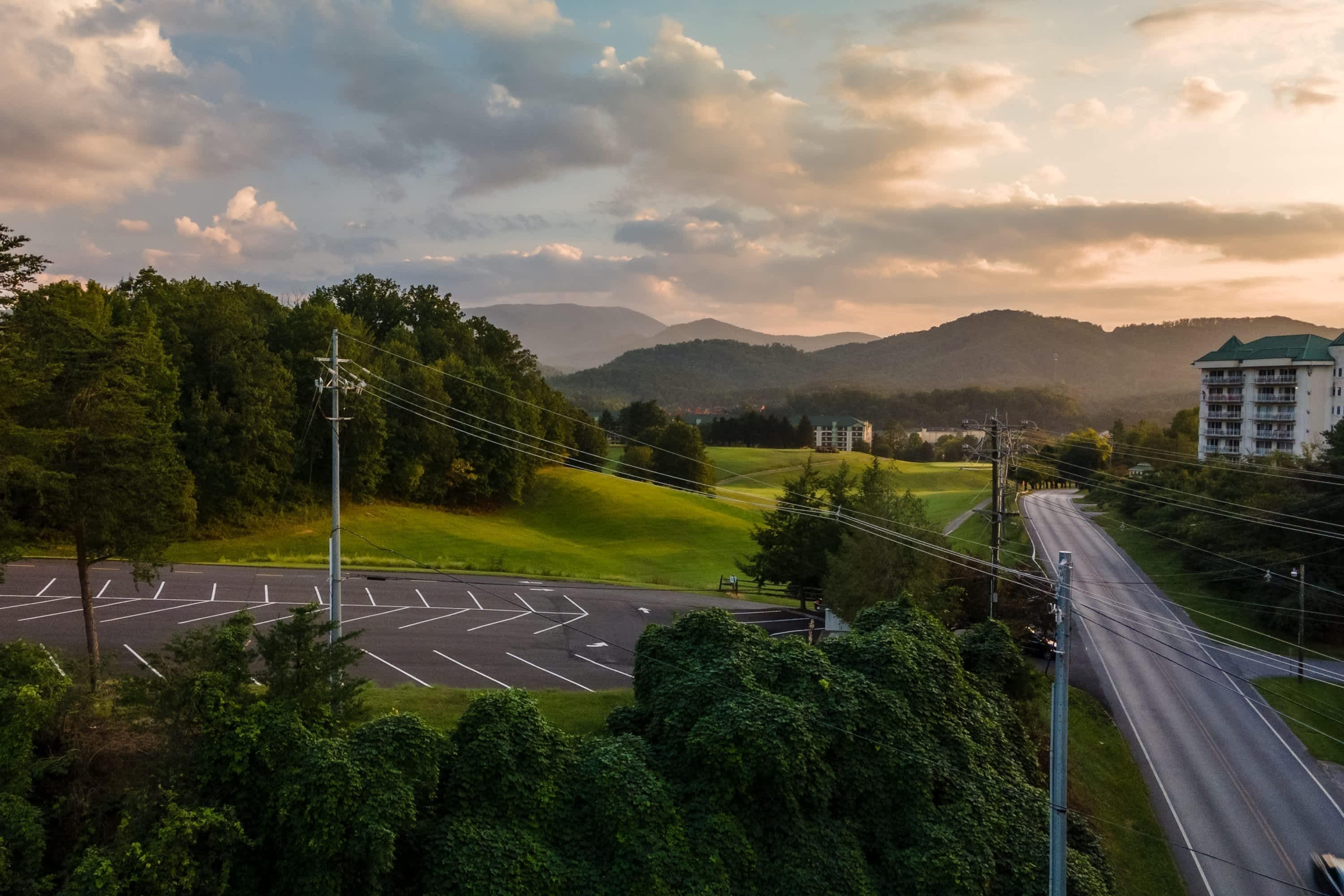 A parking lot with marked spaces, a road curving through a green landscape, and a building with a green roof in the distance.