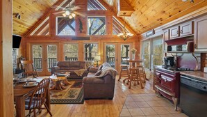 A spacious cabin interior with a dining area, living room, and kitchen, featuring wooden floors, large windows, and a high ceiling. - Cozy Mountain Retreat With Private Hot Tub (Gatlinburg)