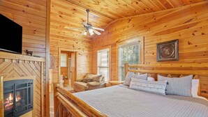 A wooden cabin interior with a fireplace, a bed, a sofa, and a ceiling fan. - Cozy Mountain Retreat With Private Hot Tub (Gatlinburg)