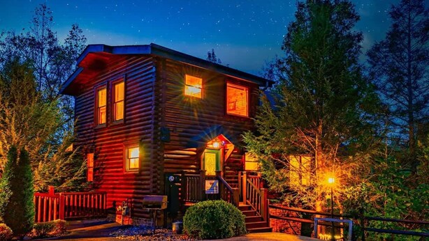 A log cabin with a porch, a staircase, and a well-lit entrance. - Cozy Mountain Retreat With Private Hot Tub (Gatlinburg)