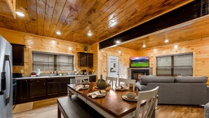 A wooden cabin interior with a dining table set for four, a gray sofa, and a kitchen area with dark cabinets and a window with blinds. - Charming New 5BR 4BA Cabin Near Dollywood (Pigeon Forge)