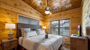 A wooden cabin bedroom with a bed, two bedside lamps, a ceiling fan, and a TV on a wooden cabinet. - Charming New 5BR 4BA Cabin Near Dollywood (Pigeon Forge)