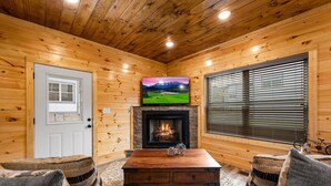 A cozy cabin interior with wooden walls, a fireplace, a television mounted above it, and a seating area with cushions. - Charming New 5BR 4BA Cabin Near Dollywood (Pigeon Forge)