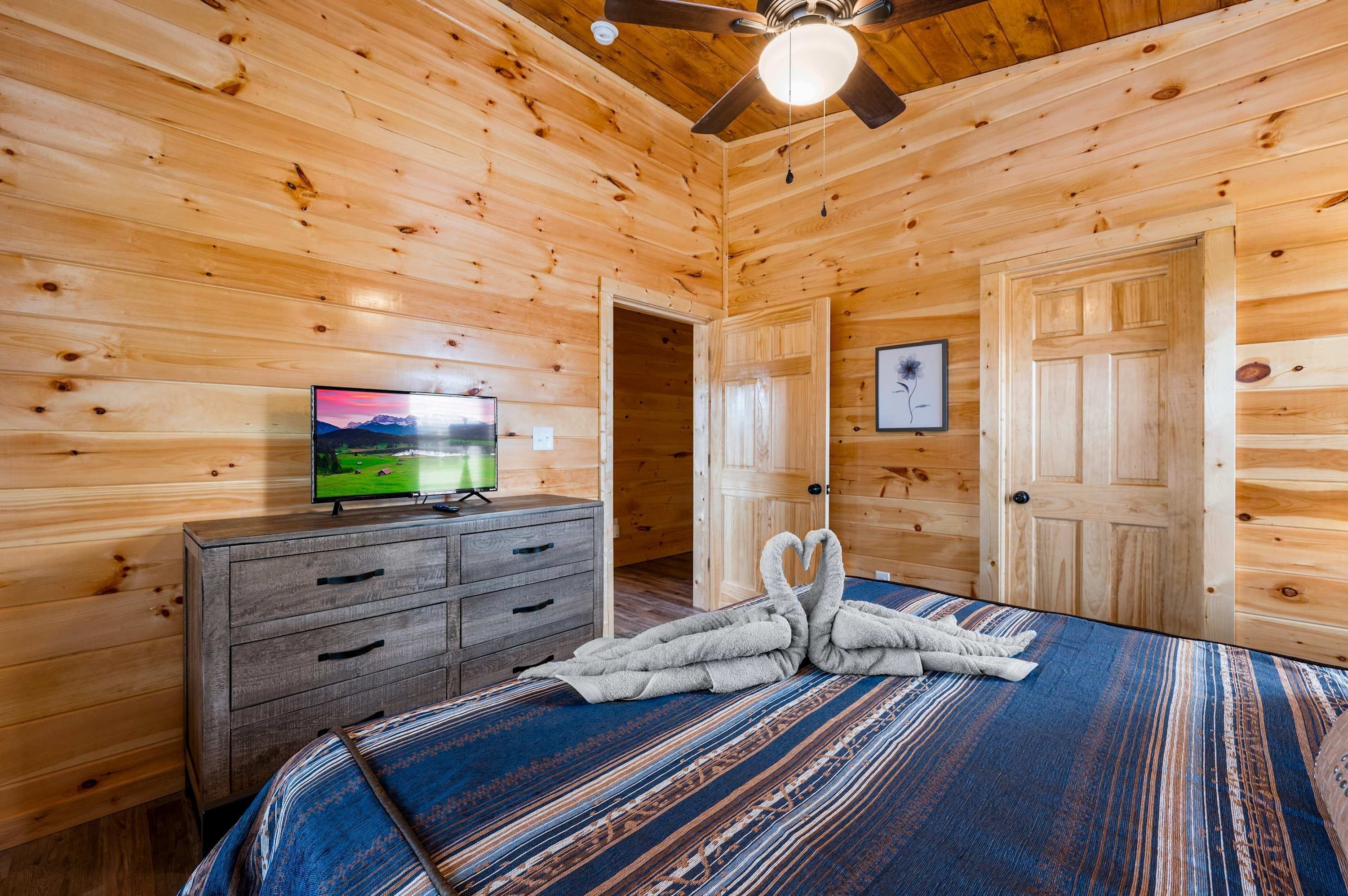 A wooden cabin room with a bed, a dresser, a television, and a ceiling fan.