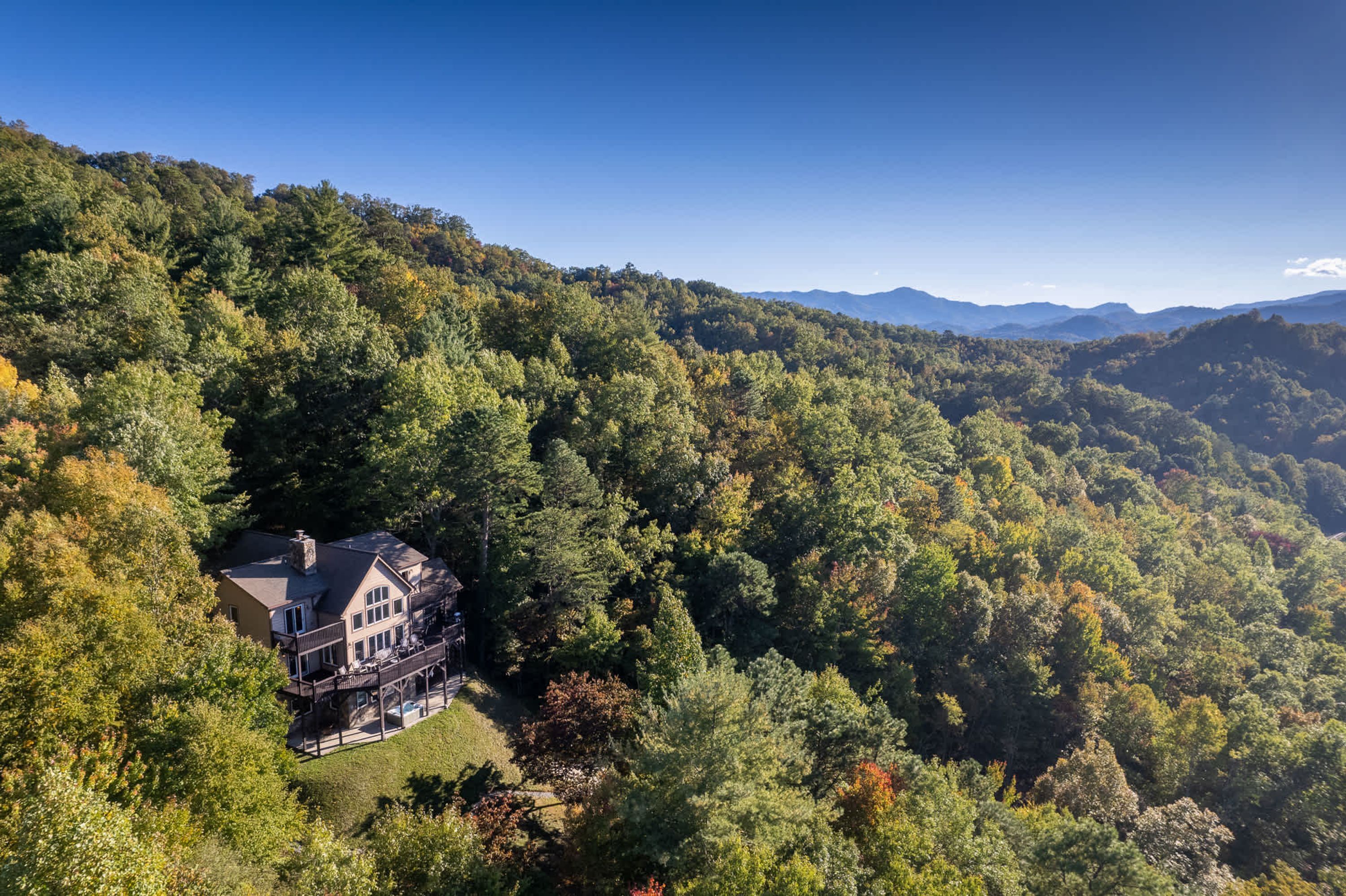 A large house with a balcony, surrounded by dense forest and mountains in the background.