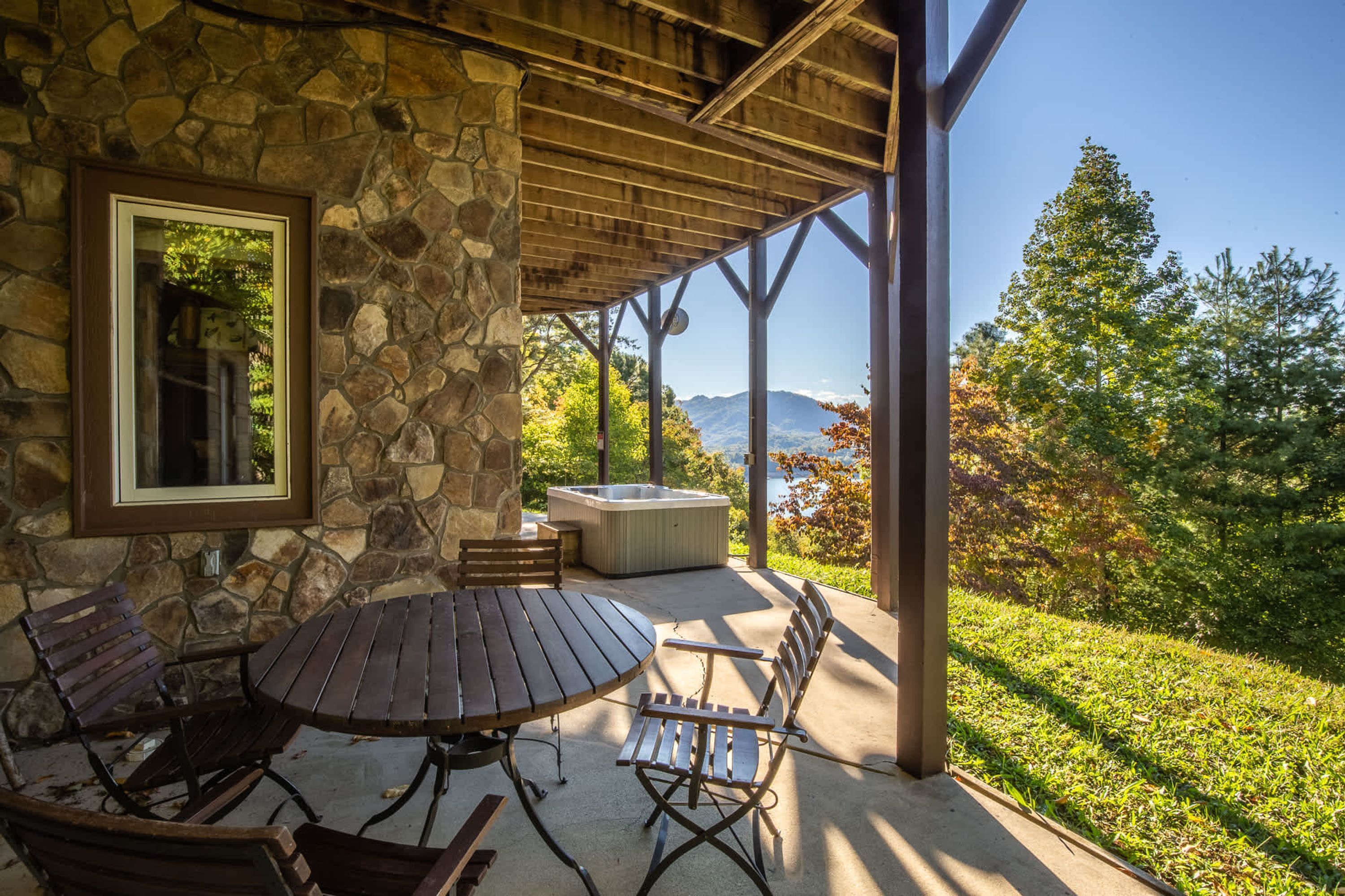Un muro exterior de piedra, una mesa de madera con sillas, un jacuzzi y vistas a montañas y árboles.