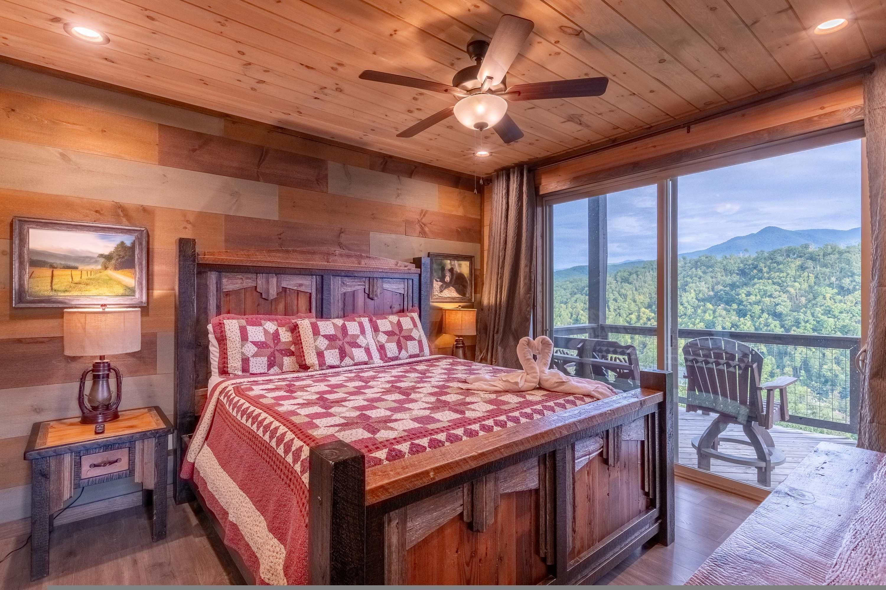 A wooden cabin bedroom with a large bed, a ceiling fan, and a balcony with a view of the forest.