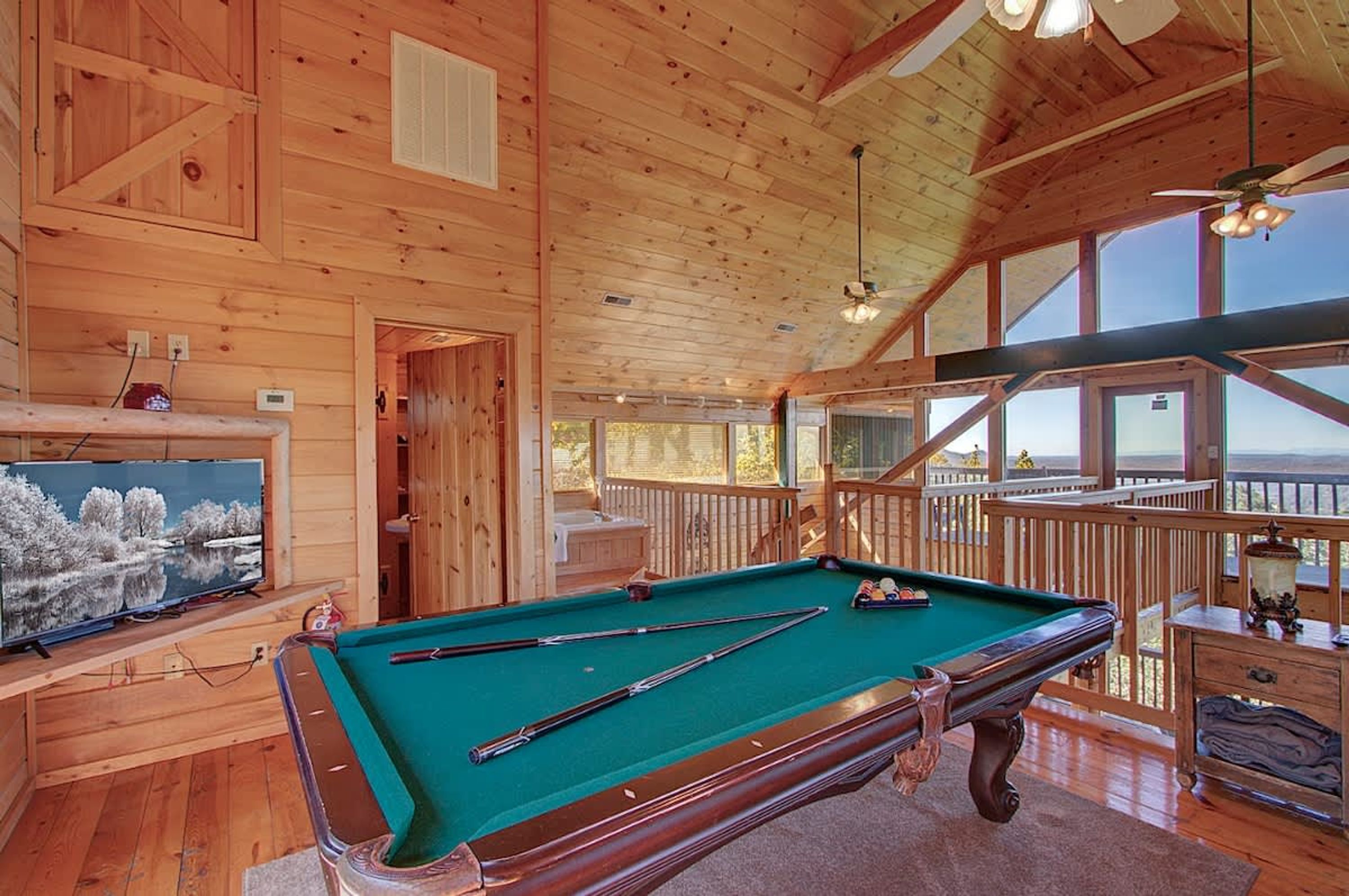 A wooden interior with a pool table, a television, and a balcony with a view.