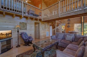 Eine geräumige Holzhütte mit einem Balkon, einem gemütlichen Wohnbereich mit einer Couch und einem Couchtisch sowie einem Fernseher auf einem Standfuß.