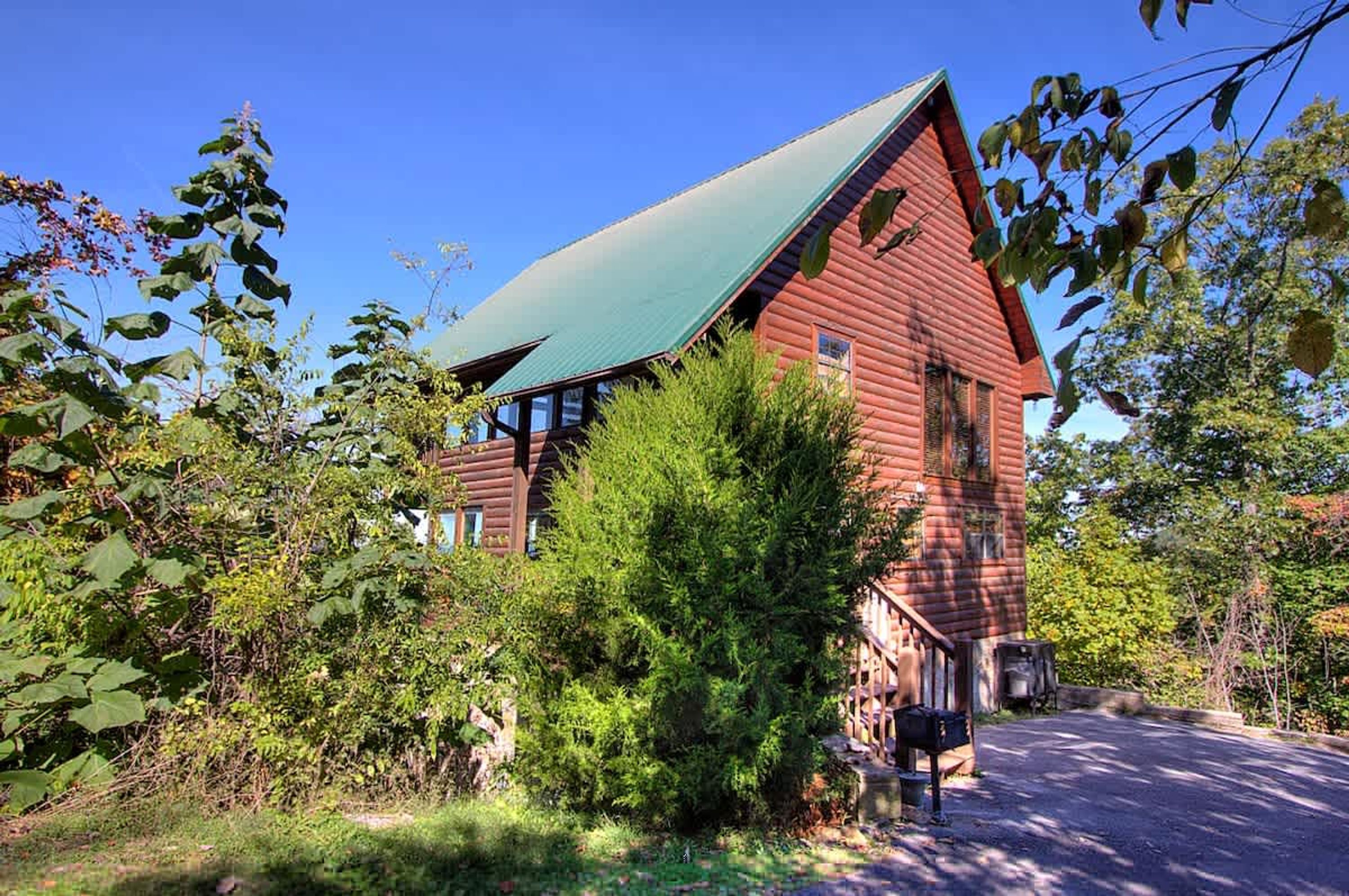 A rustic log cabin with a green roof, surrounded by dense greenery and a clear blue sky.