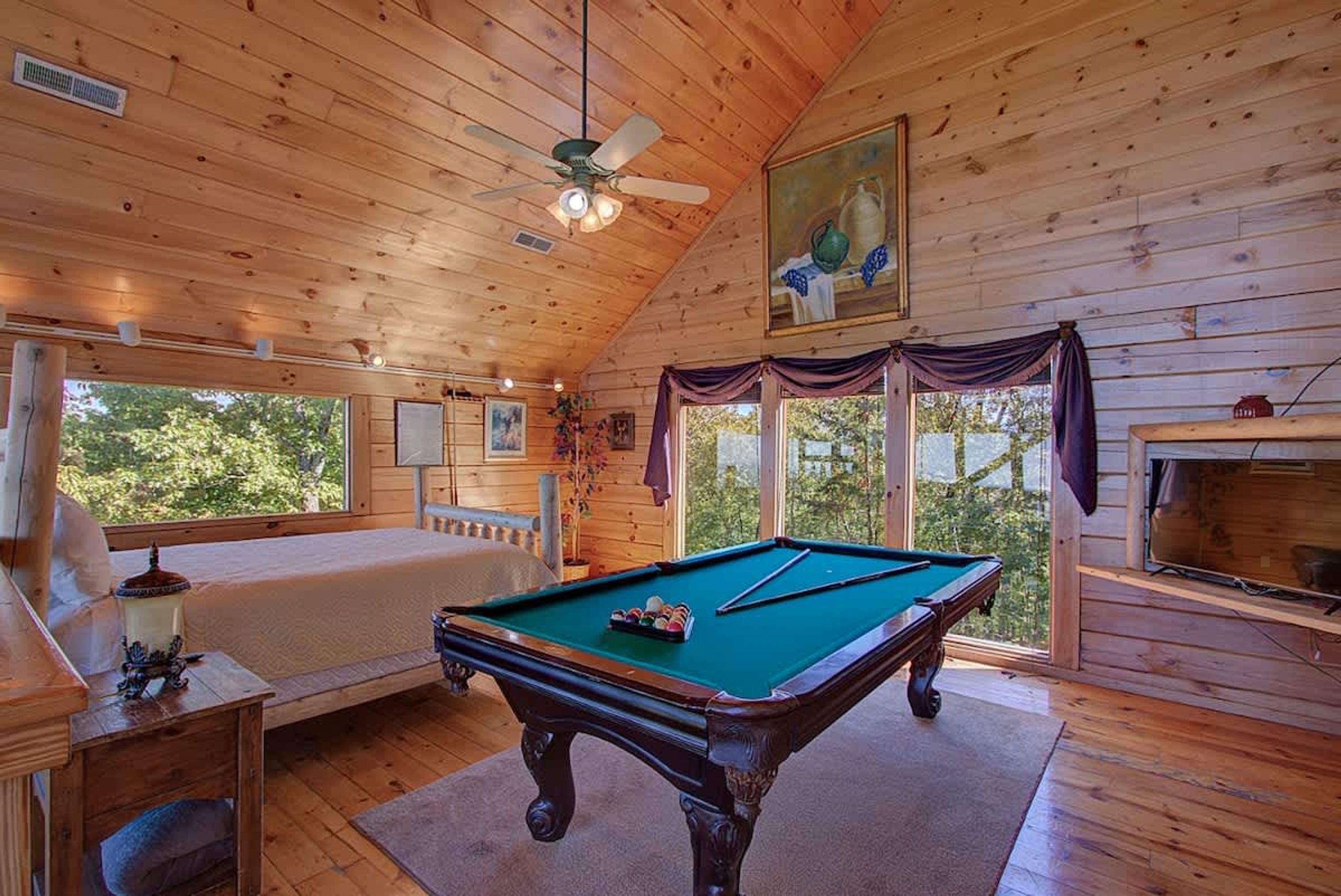 Een houten kamer met een pooltafel, een plafondventilator en een groot raam met uitzicht op bomen.