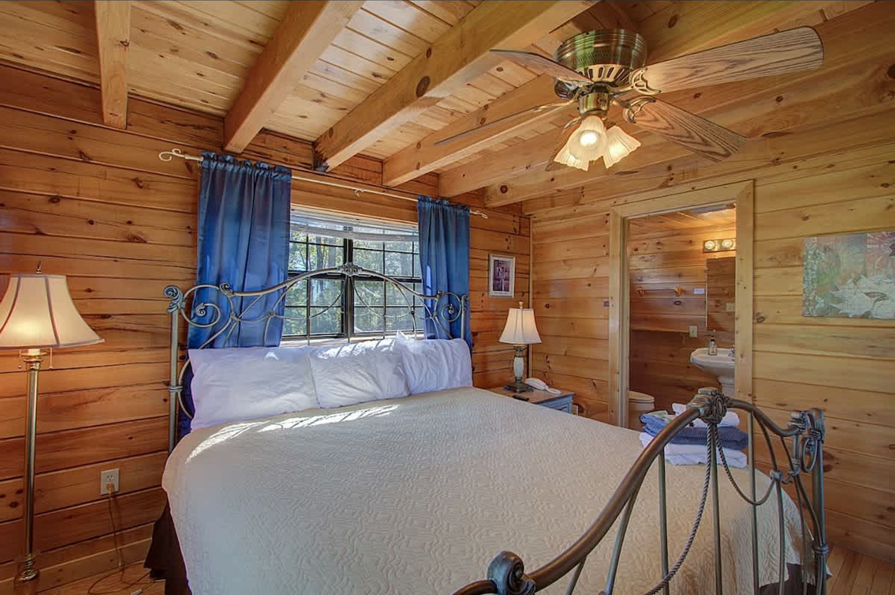 A wooden cabin bedroom with a bed, two bedside lamps, a ceiling fan, and a window with curtains.