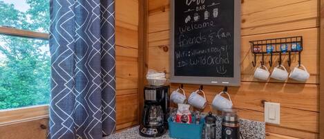 Een knus interieur met een koffiebar, een krijtbordmenu en een raam met uitzicht op bomen.
