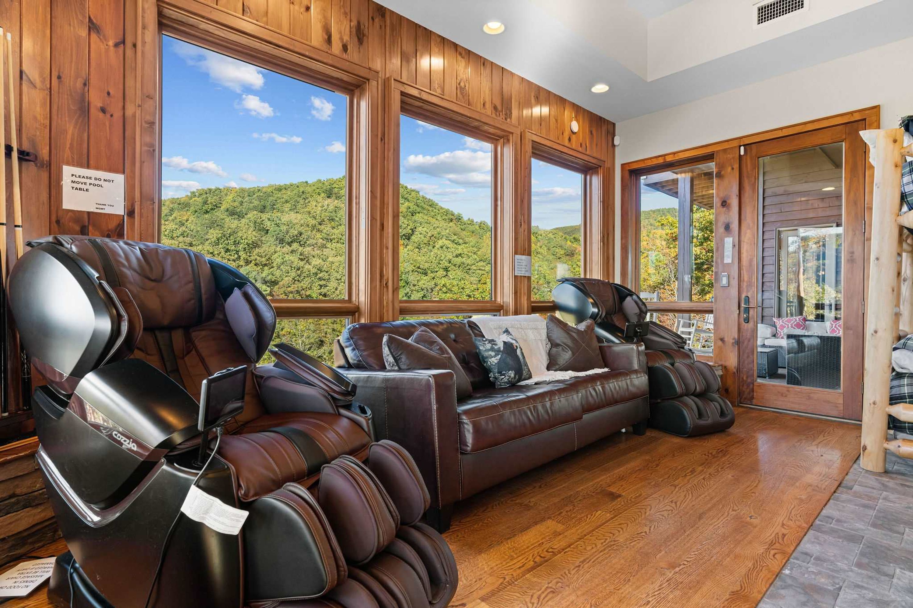 A massage chair and a sofa in a room with large windows overlooking a forested hill.