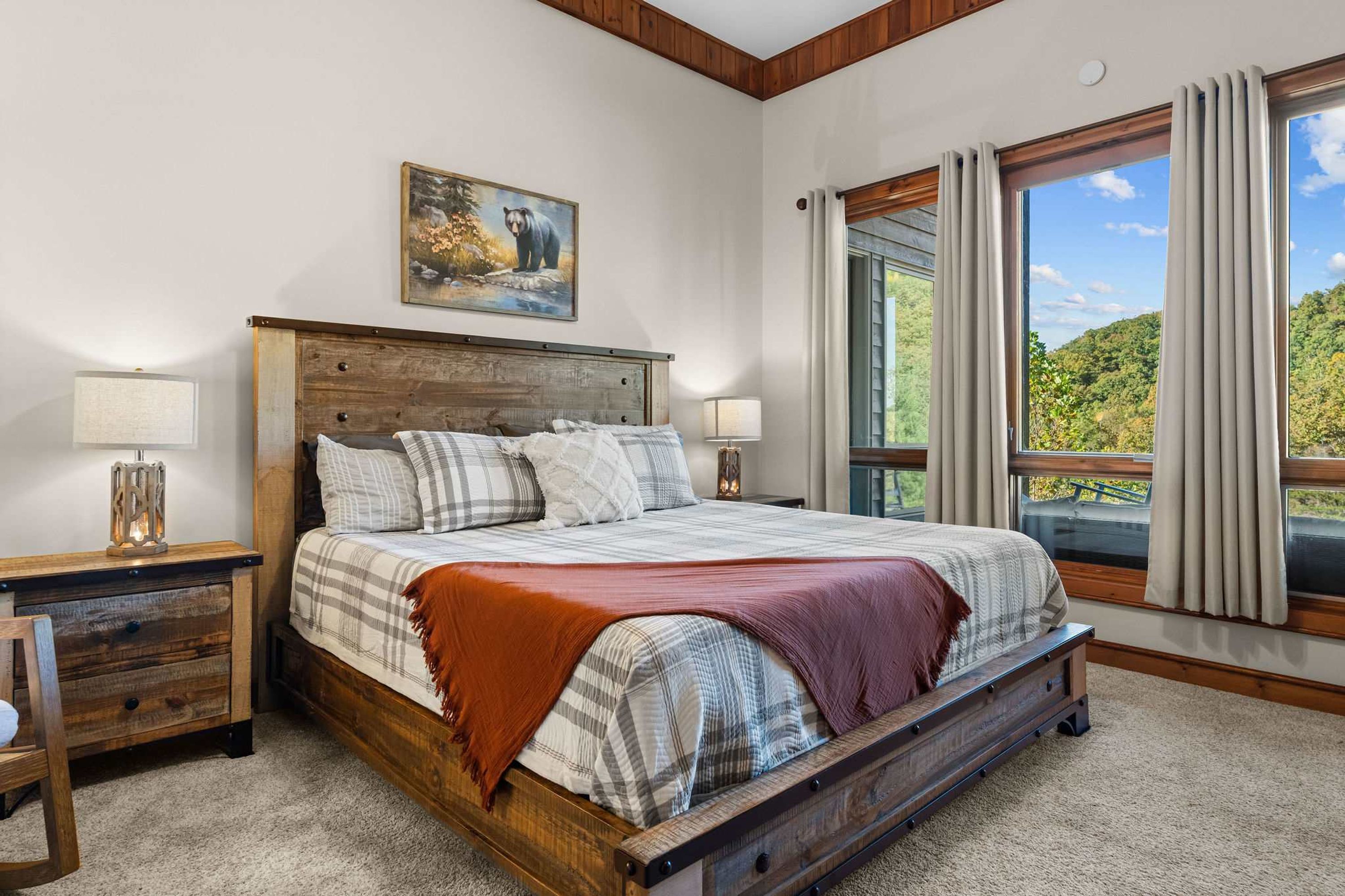 Un dormitorio con una cama de madera, dos mesitas de noche, una lámpara auxiliar, un cuadro y un ventanal con cortinas.