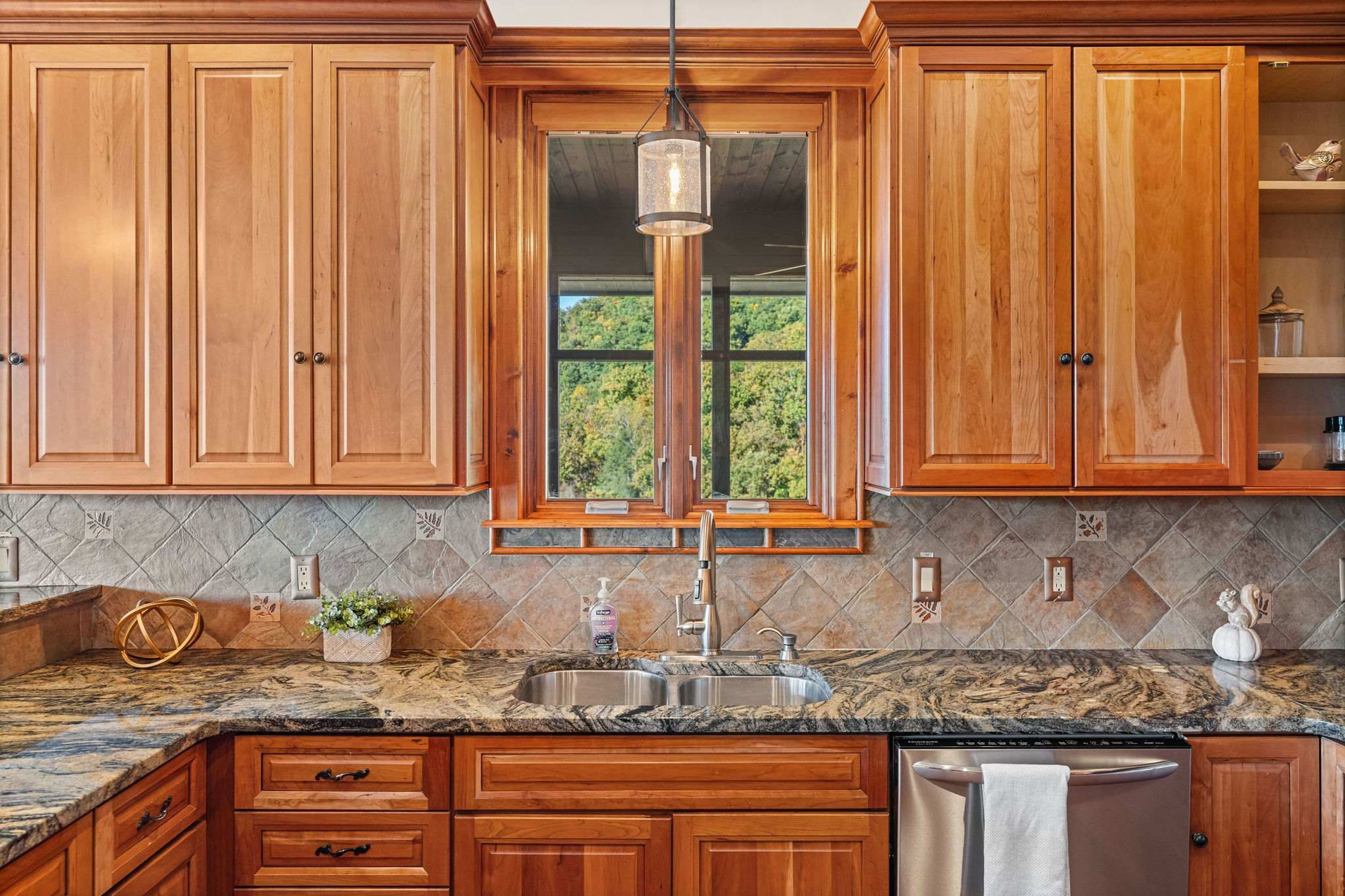 A kitchen with wooden cabinets, a granite countertop, a stainless steel sink, and a window with a view of greenery.
