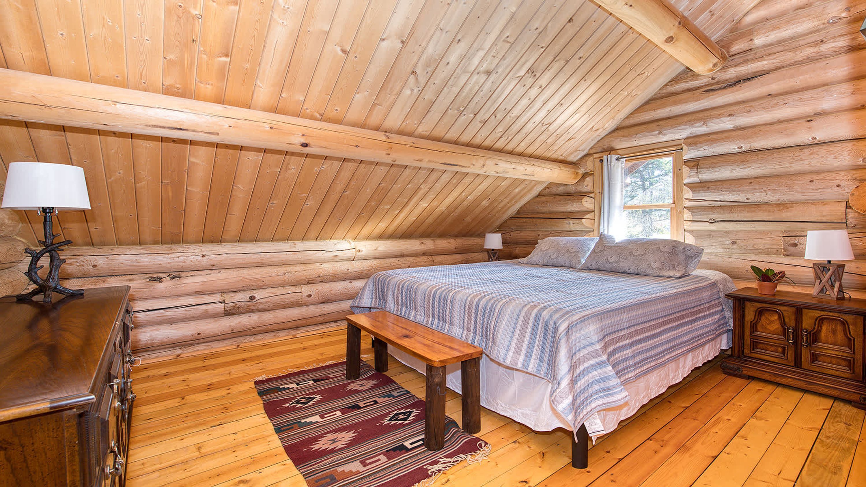 Une chambre située dans un chalet en rondins, avec un lit, un banc, des tables de chevet et une fenêtre.
